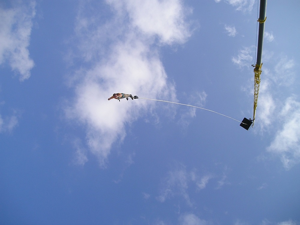 Bungee Jumping: A bungee jumper is falling toward the ground after jumping from a crane. Five spectators or instructors stand along the crane beam.